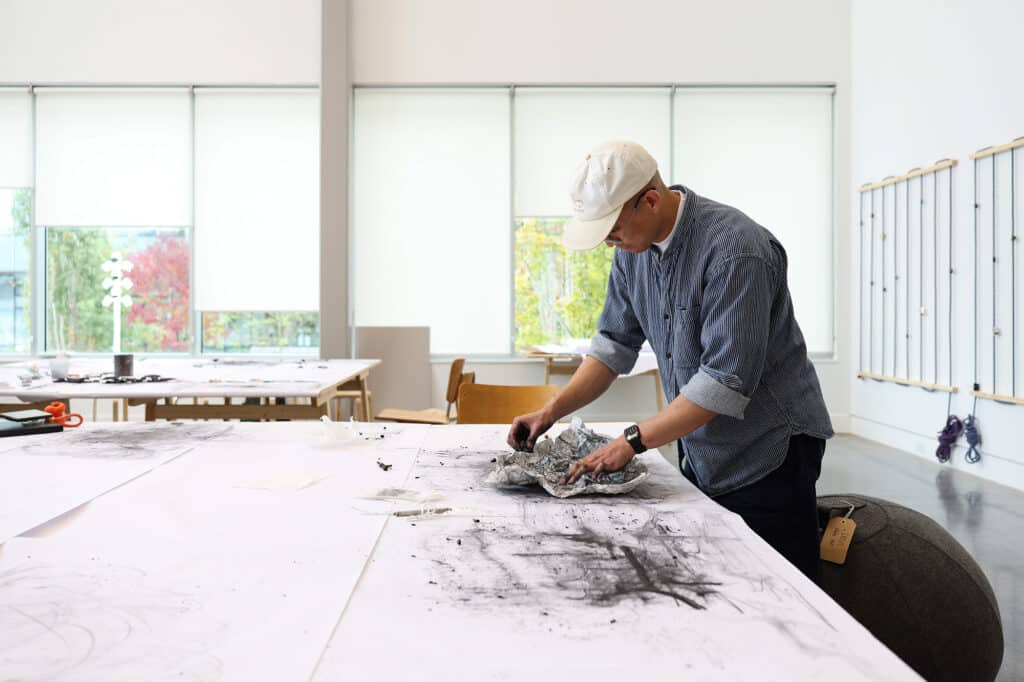 A person in a white cap works with charcoal-covered paper at a large table in a bright art studio.