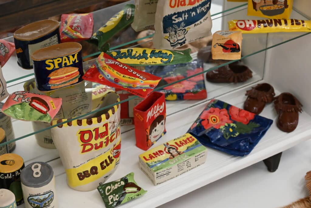 Colourful ceramic sculptures resembling vintage food packaging, including Spam, Old Dutch chips and candy bars, displayed on glass shelves alongside ceramic shoes.