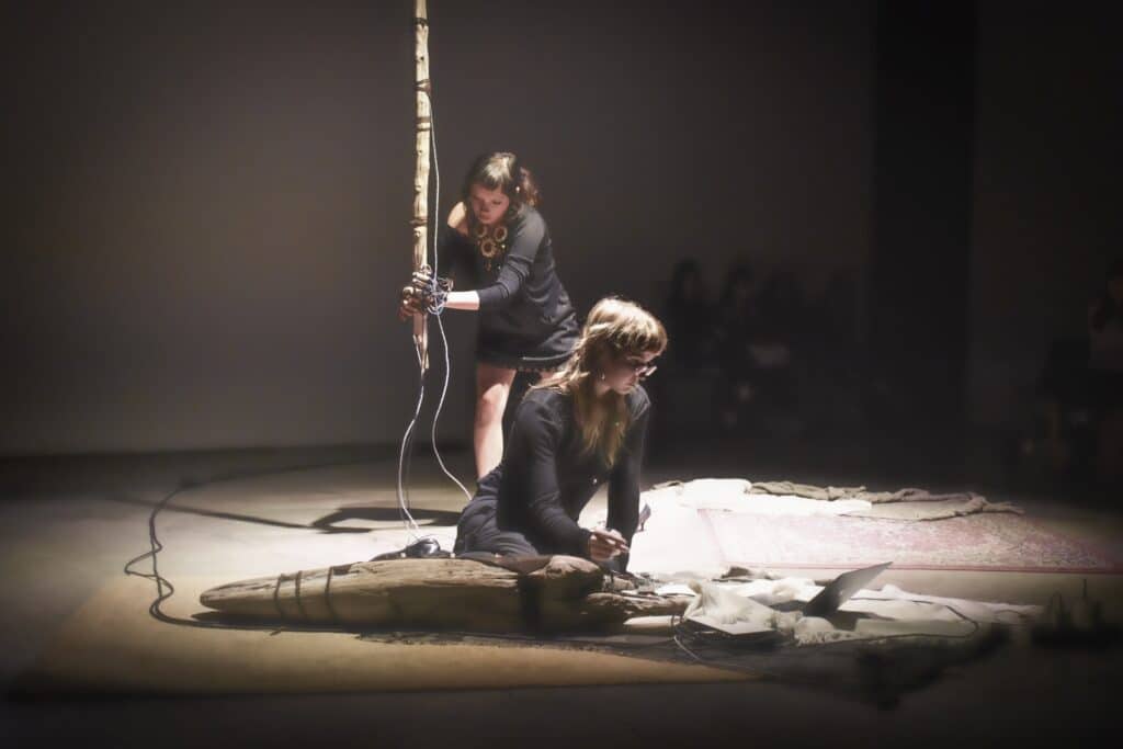 Two performers work with an interactive installation under spotlights. One kneels beside a laptop and electronic equipment while the other holds a suspended wooden object.