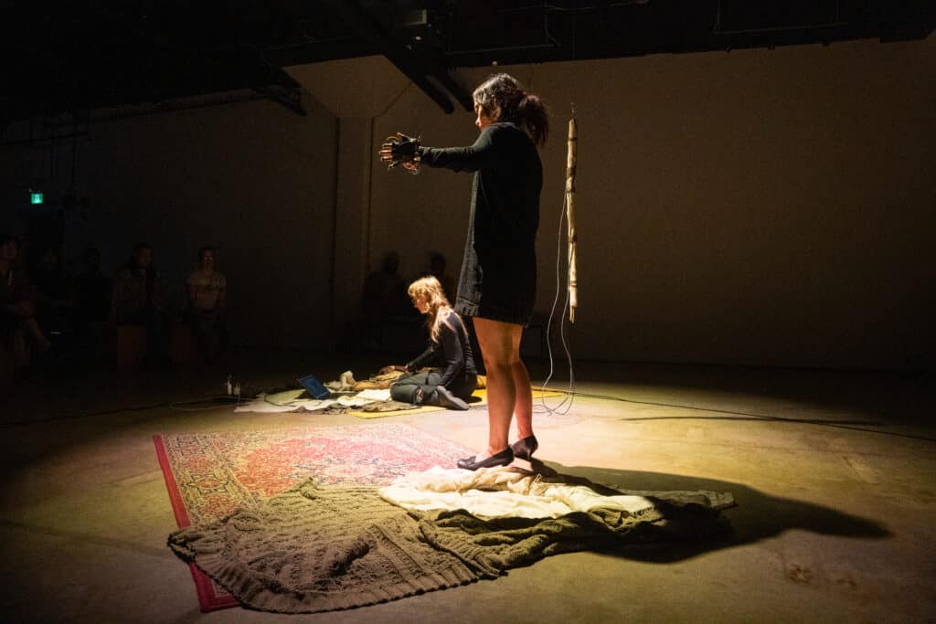 A performer stands under a spotlight with arms extended, wearing wired devices on their hands, while another performer sits on the floor near a laptop. Rugs and fabric cover the performance space as an audience watches from the shadows.