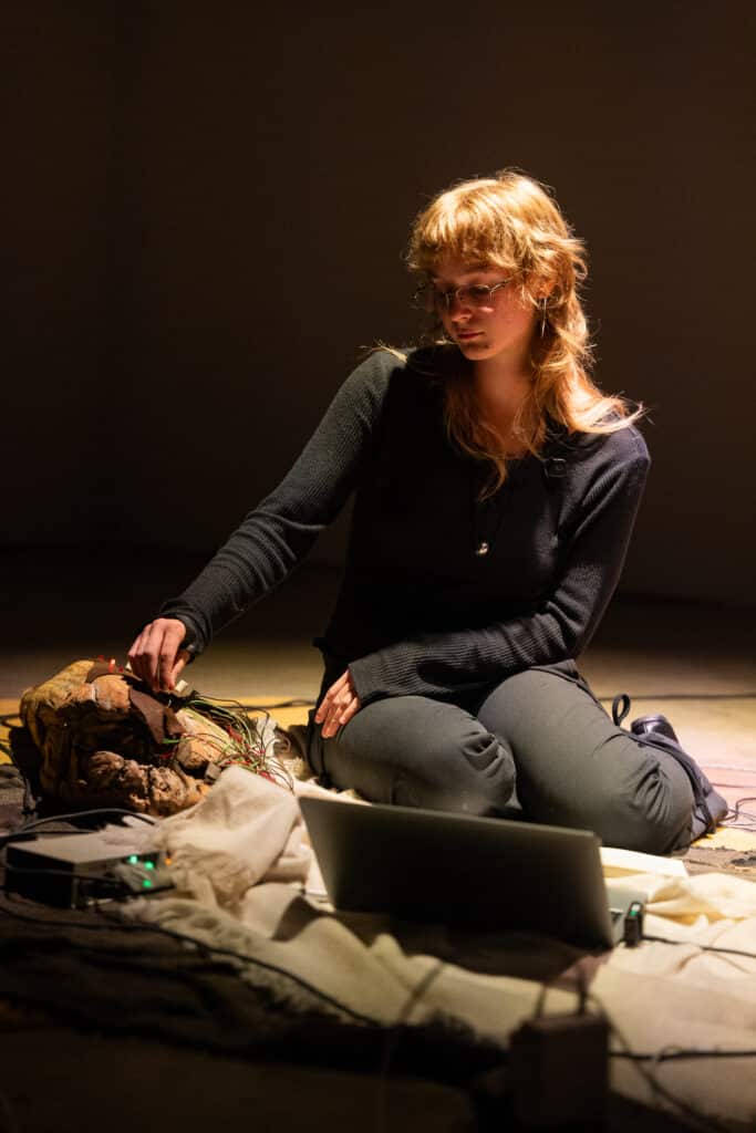 A performer kneels under a spotlight, adjusting wires connected to a piece of driftwood beside a laptop and electronic equipment.
