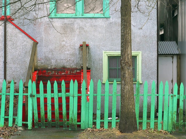 A bright green picket fence and red steps stand in front of a grey building with green-trimmed windows and a tree in the foreground.
