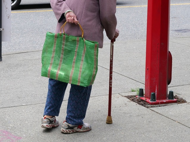 Street photograph focused on a person carrying a large green striped bag and using a cane beside a red post.