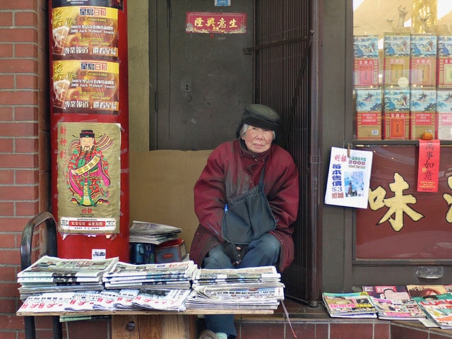 A person sits behind stacks of newspapers outside a storefront surrounded by colourful signs and posters.