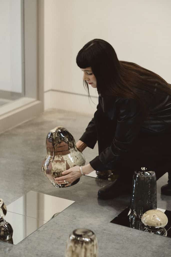 An artist crouches to arrange a glazed ceramic sculpture on a mirrored surface, surrounded by other ceramic works in a gallery.