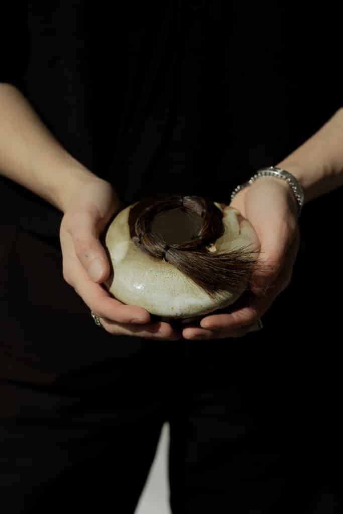 A person dressed in black holds a rounded, cream-coloured ceramic vessel adorned with a coiled bundle of dark hair.