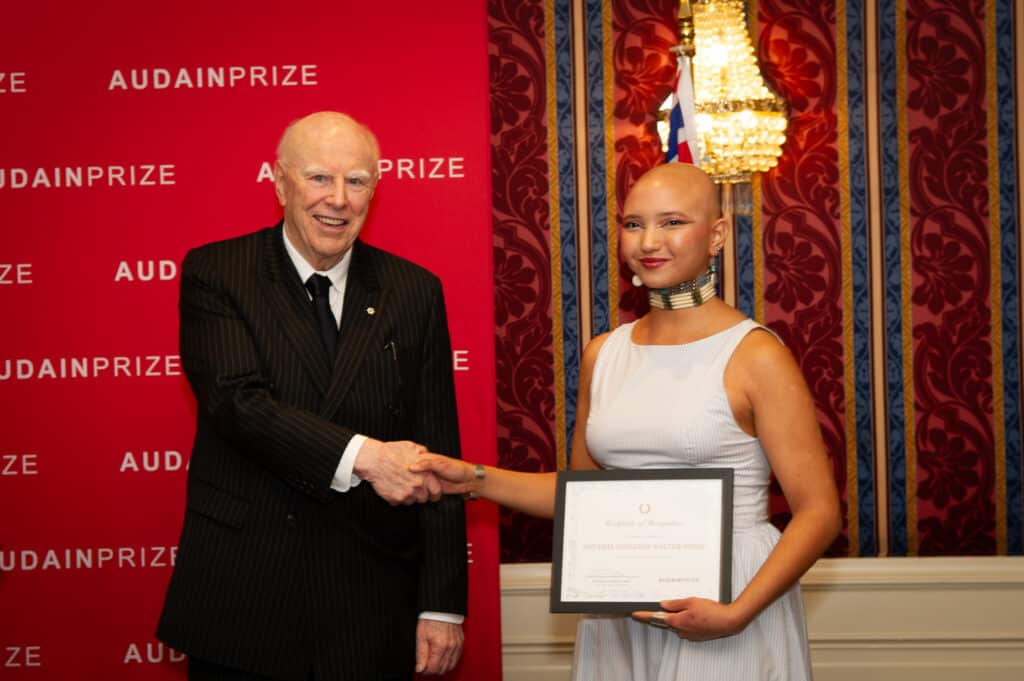 An older person in a black pinstripe suit shakes hands with artist Natasha Ashleigh Walter-Pinay, who holds a certificate of recognition in front of a red Audain Prize backdrop.