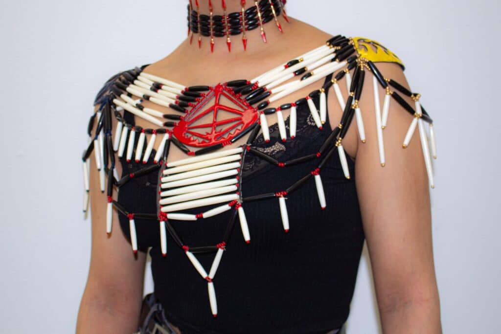 Close-up of a person wearing an elaborate beaded necklace and shoulder adornment featuring black and white tubular beads, red accents and a geometric red centrepiece over a black top.