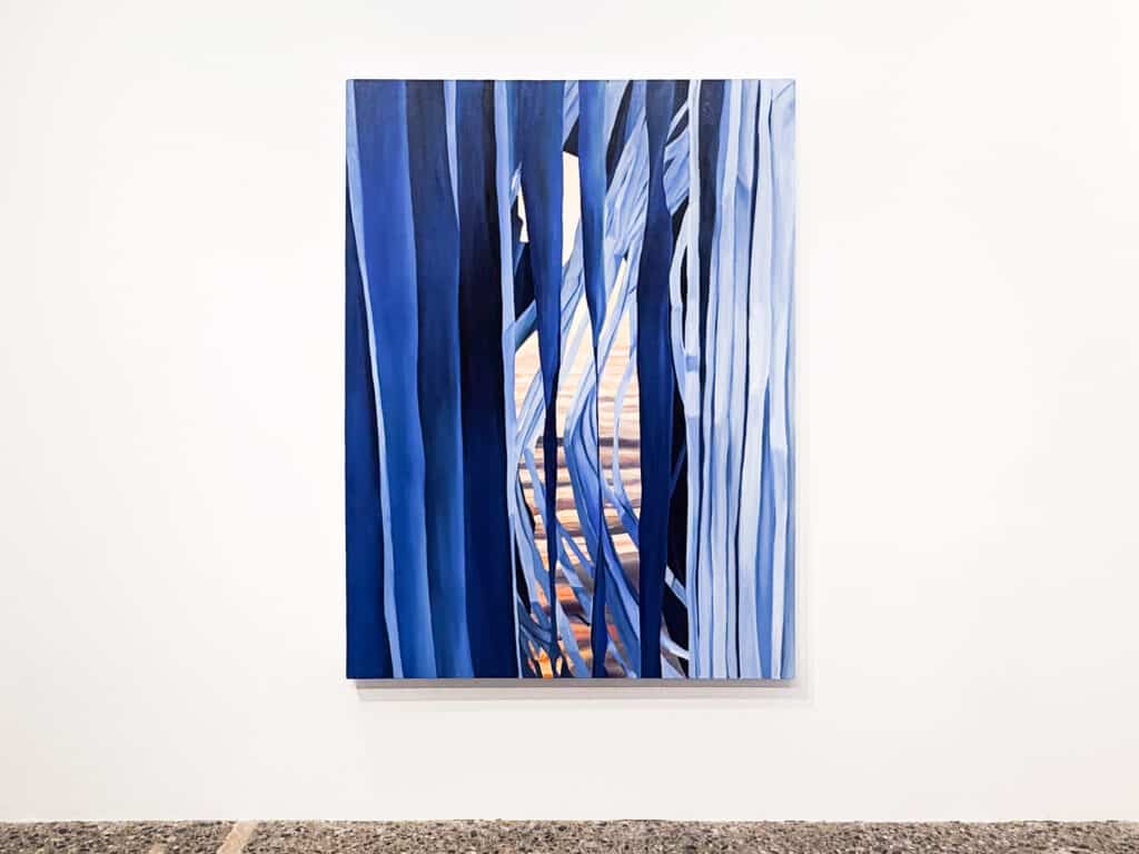 A vertical painting of layered, flowing blue forms in shades of navy and pale blue, revealing glimpses of a warm orange and pink landscape, hangs on a white gallery wall.