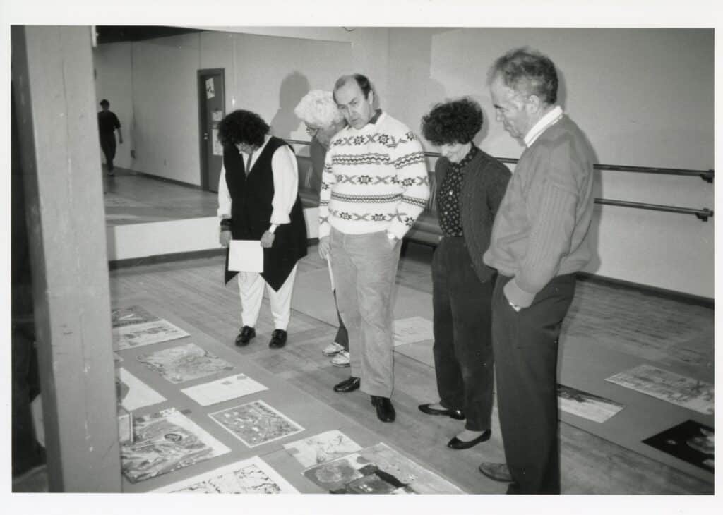 Black-and-white archival photograph of five people standing together, examining artworks arranged across the floor of a large room.