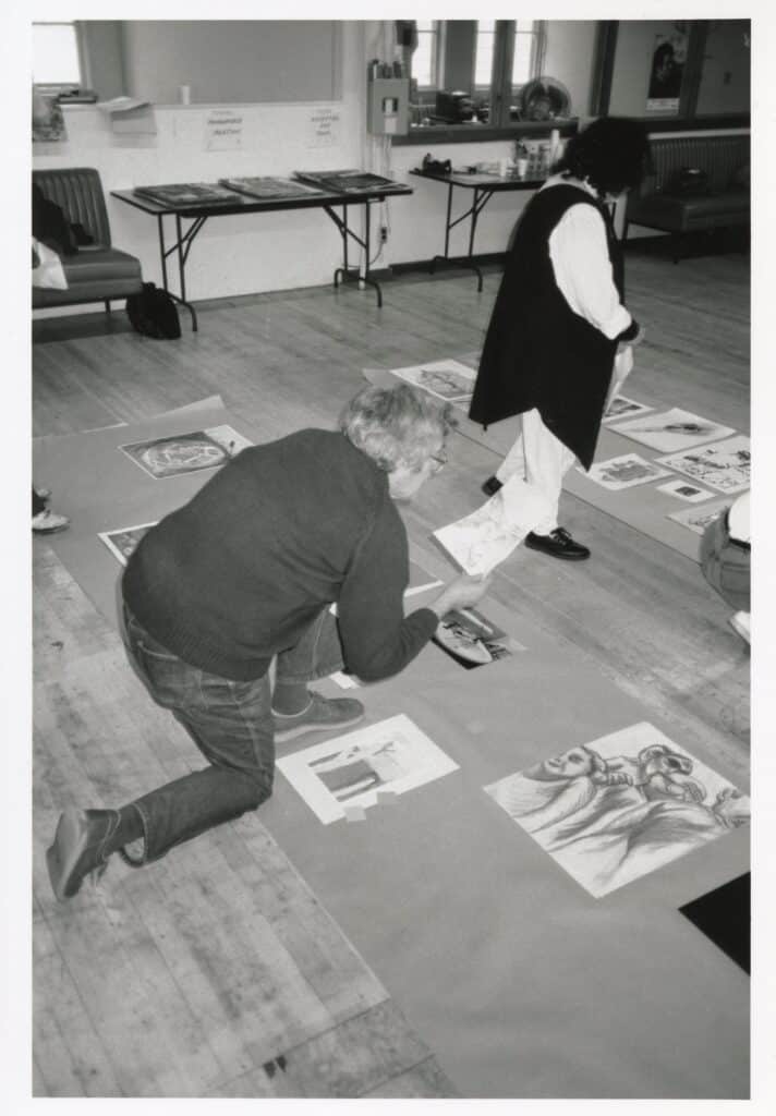 Black-and-white photograph of two people examining artworks laid out on the floor, one kneeling to lift a drawing while the other stands nearby.