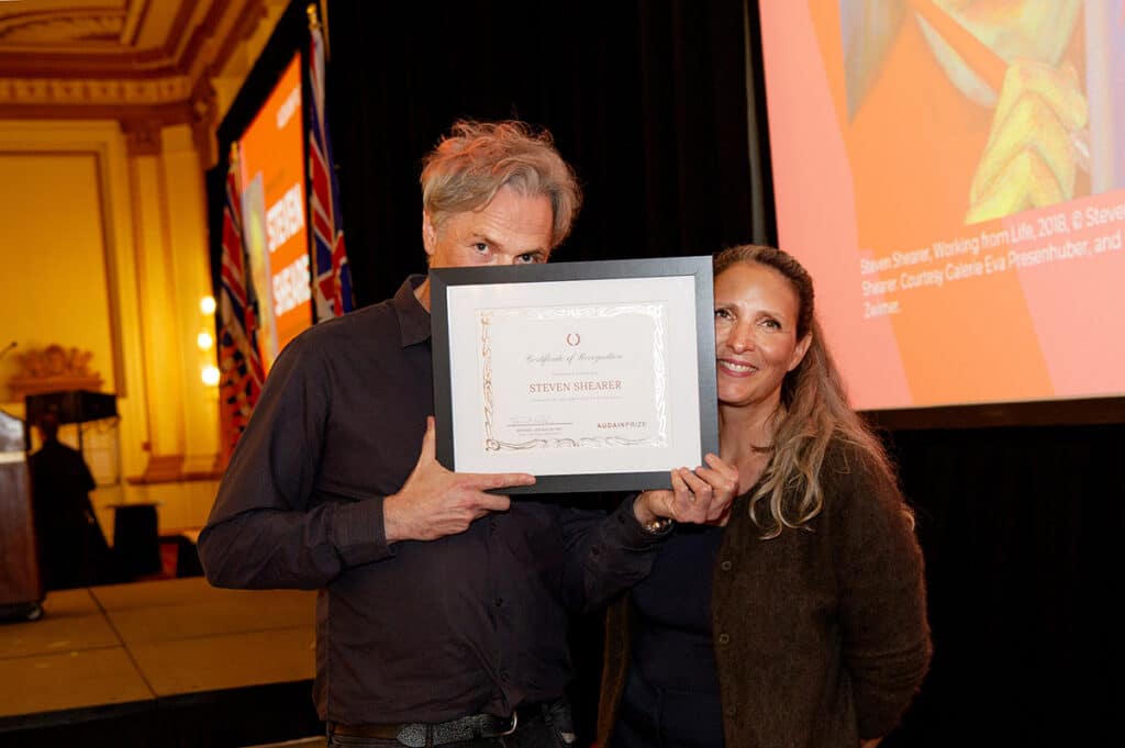 Two people pose at an awards ceremony, holding a framed certificate recognizing artist Steven Shearer, with a projection of artwork behind them.