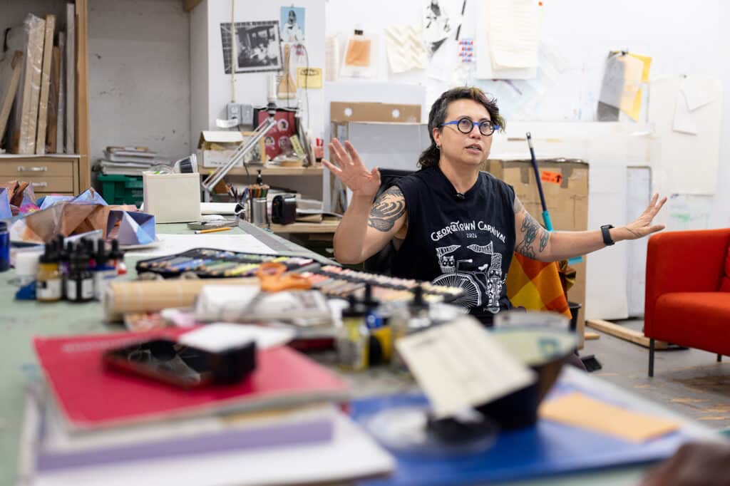 A person sits in an artist's studio speaking with both hands raised, surrounded by works in progress and art supplies.