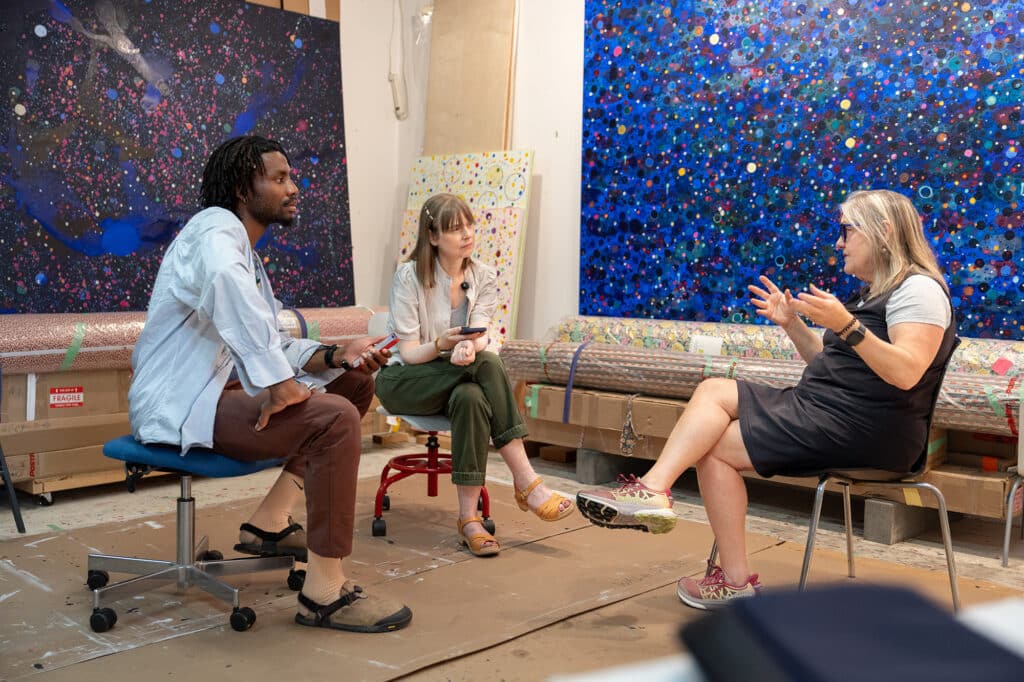 Two interviewers seated across from an artist in a studio as the artist gestures while speaking. Large abstract paintings surround the group.