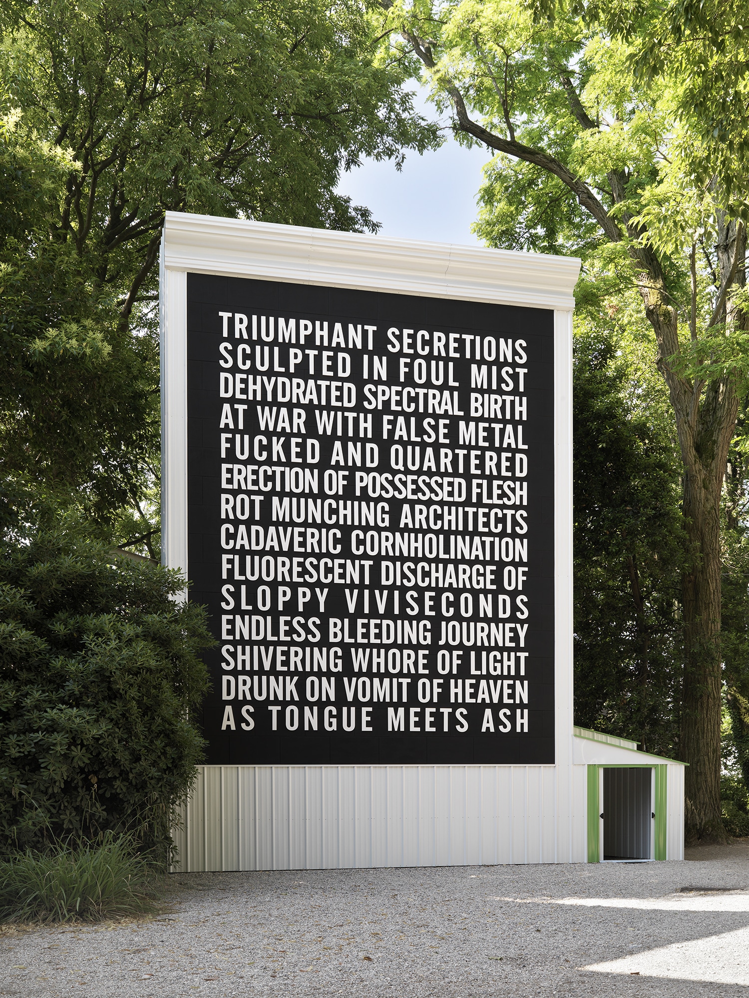 A large black-and-white text installation on a white building surrounded by leafy trees, featuring a poem in bold capital letters beginning with “Triumphant Secretions.”