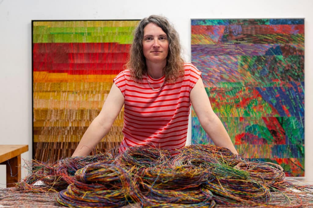 A person with long hair and striped red shirt stands smiling above a table covered in telecommunications wires with a pair of brightly coloured artworks in the background.